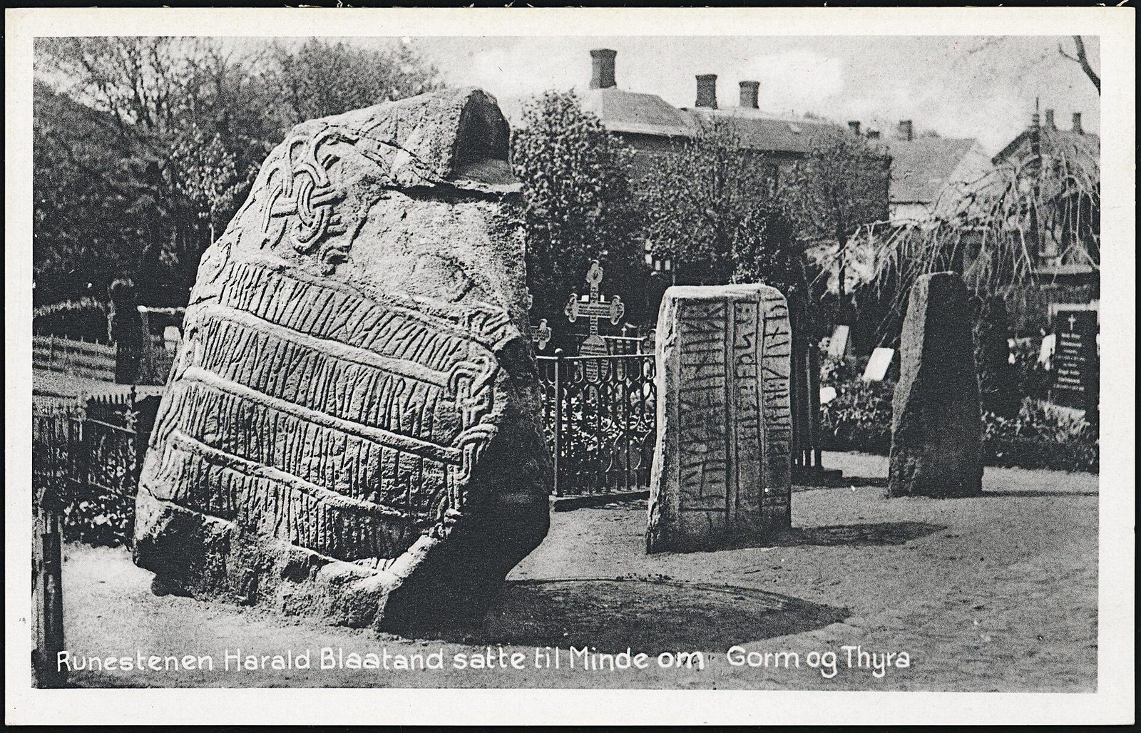 A black-and-white photograph of a large carved boulder in a churchyard, with a smaller standing stone behind it.