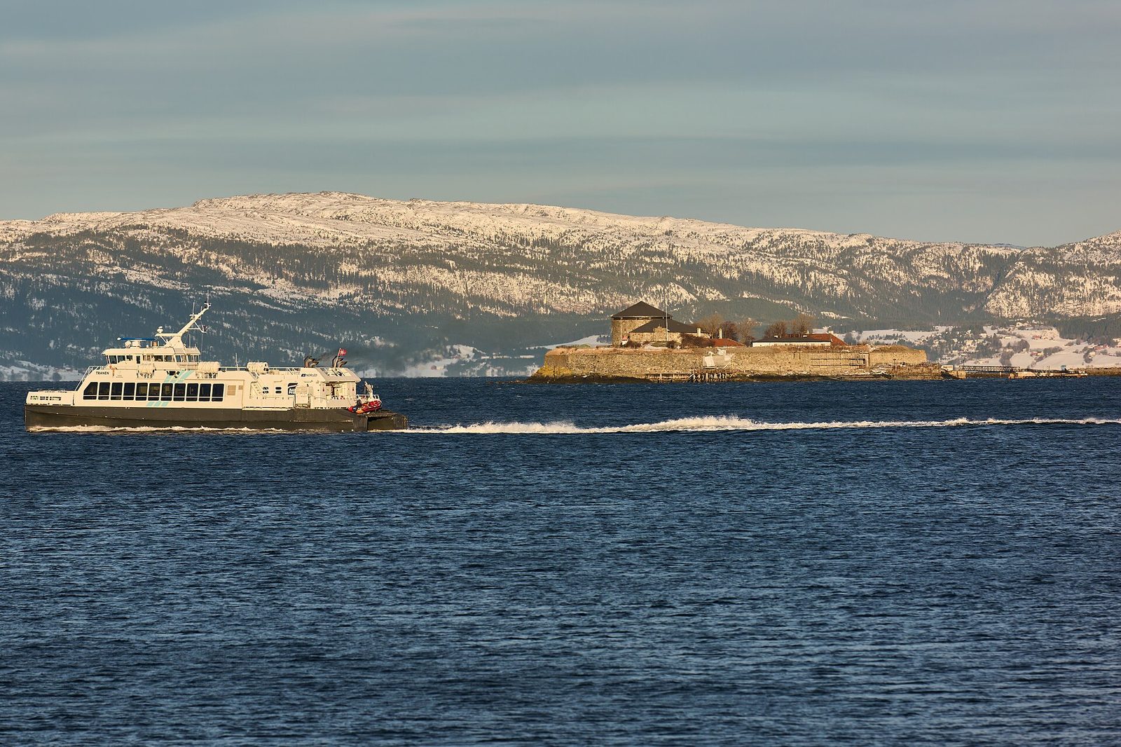 A small fortified island in a fjord, a passenger ferry passing it, snow on the hills behind.