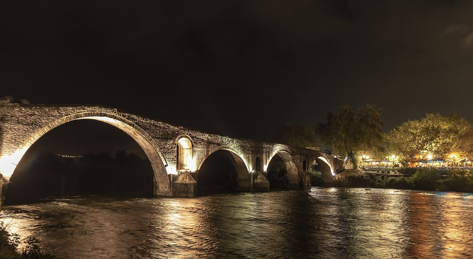 A stone bridge of four unequal arches crossing a wide green river, the largest arch rising to a sharp point at the centre, with wooded hills behind.