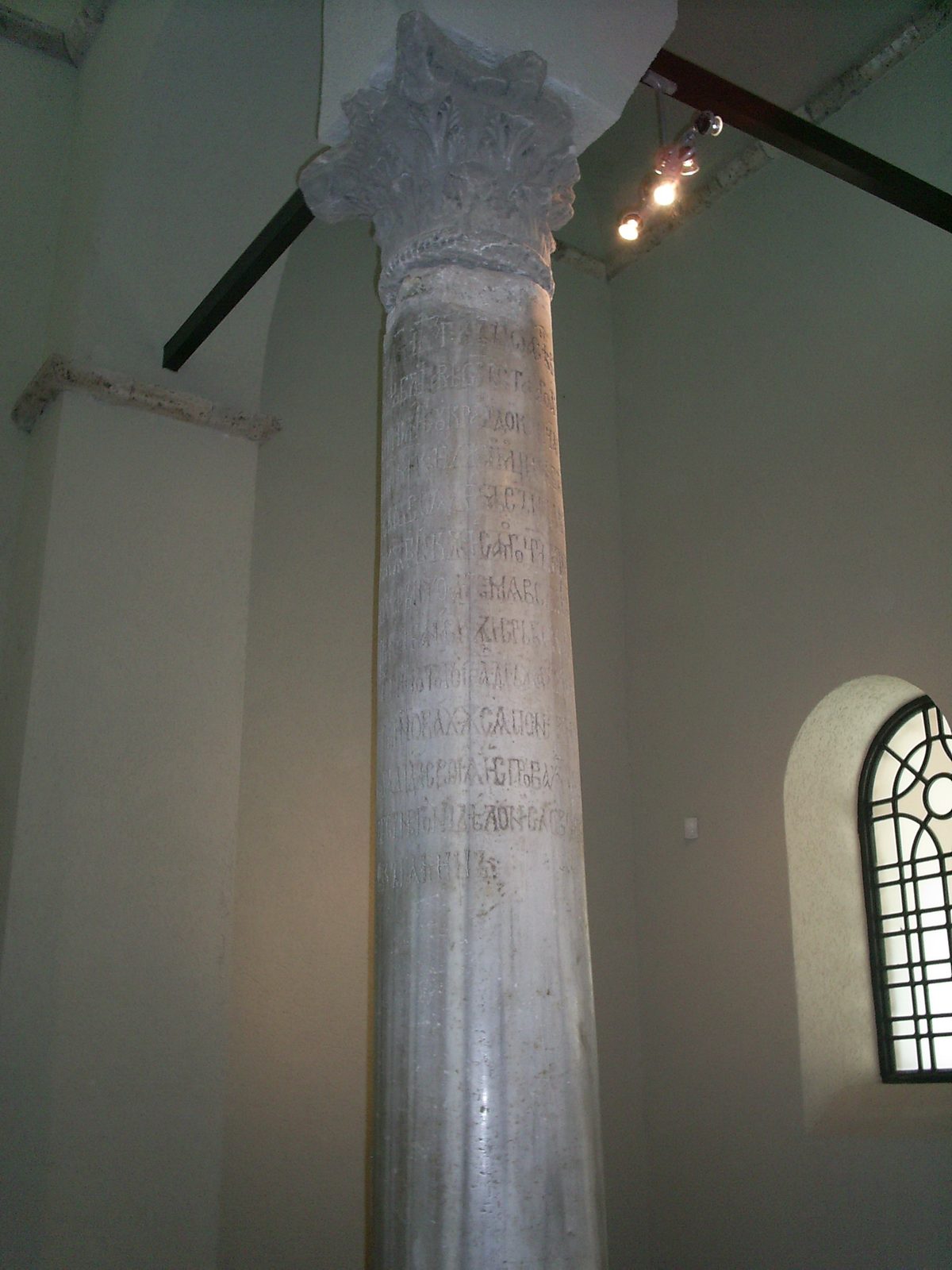 A tall slender stone column standing inside a church, its shaft carrying a long inscription in closely cut Cyrillic letters running round and down it.