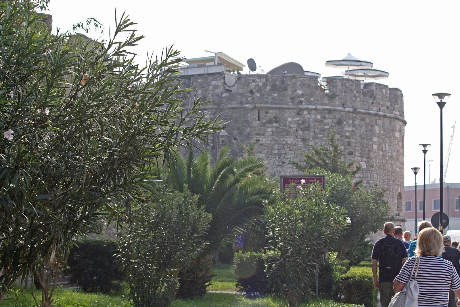 A squat round tower of rough stone and brick standing at the end of a stretch of old city wall, with modern buildings and a paved square around it.