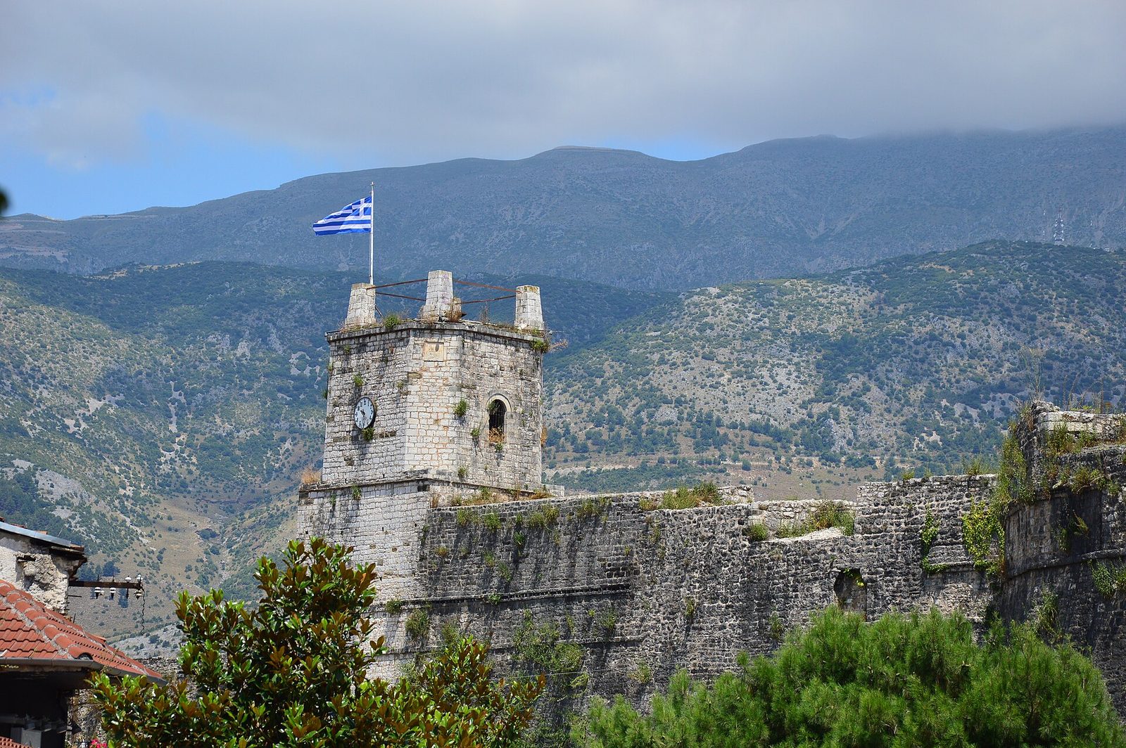 A long fortified wall with round and square towers following the edge of a lakeside promontory, a town of tiled roofs behind it and still water in front.