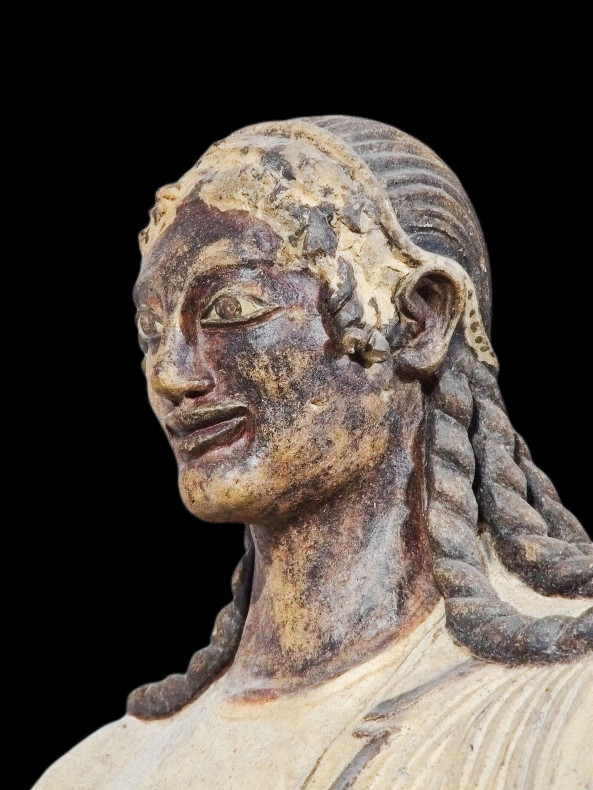 A painted terracotta head of a smiling god with long braided hair, against a black ground.