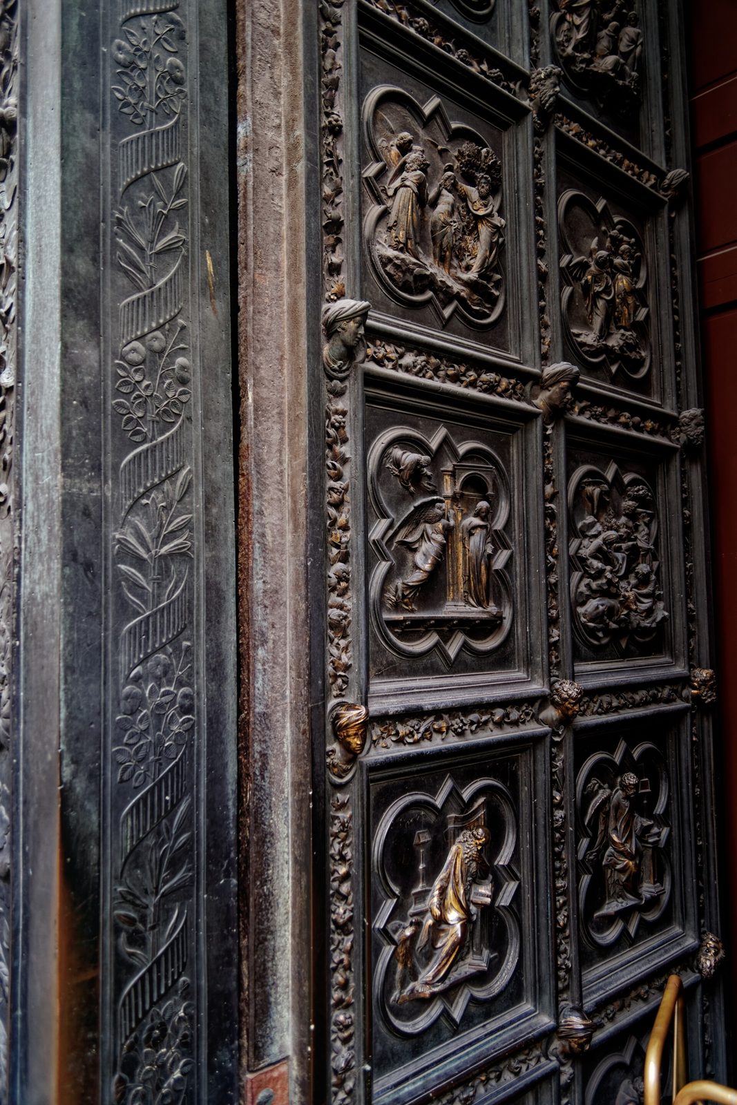 A pair of dark bronze doors seen at a sharp angle, divided into rows of small relief panels, each scene framed inside a four-lobed moulding set against a patterned ground.