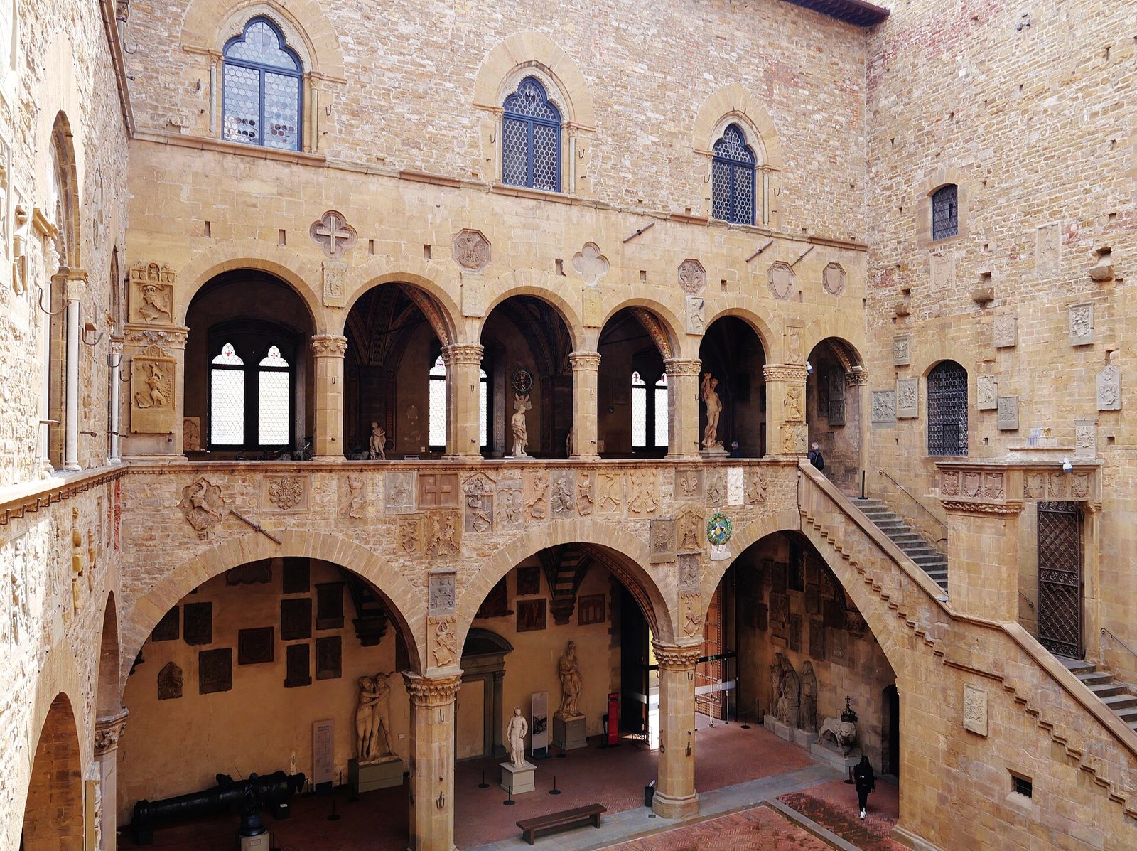 A courtyard enclosed by rough stone walls, with a vaulted arcade at ground level, an open loggia of pointed arches above it and a covered stone stair climbing one side; carved coats of arms are set into the masonry.