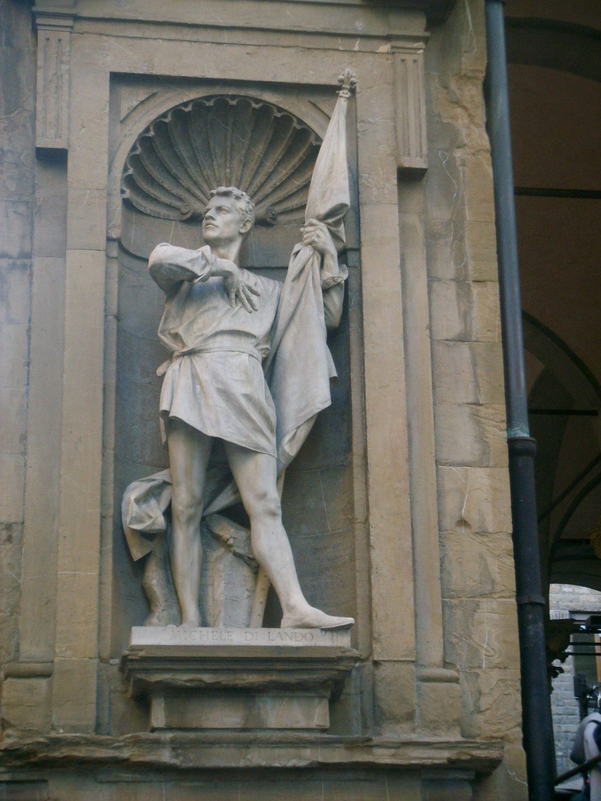 A marble statue in a shallow niche: a man in a short belted tunic and hose, bare-legged, standing with one arm raised to hold a furled banner against his shoulder.