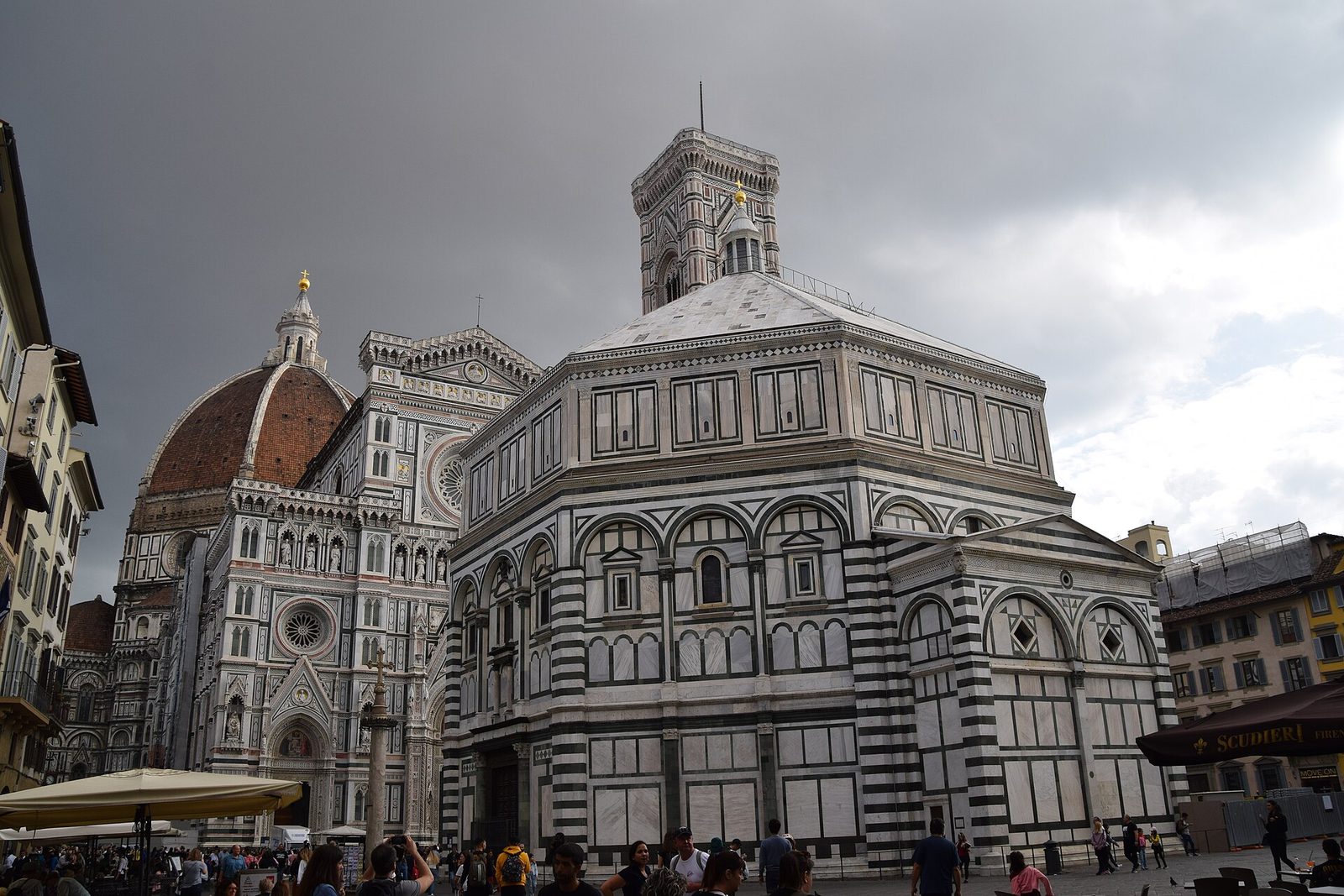 An octagonal building faced in green and white marble panels, standing in a paved square with a much larger striped cathedral and a tiled dome rising behind it.