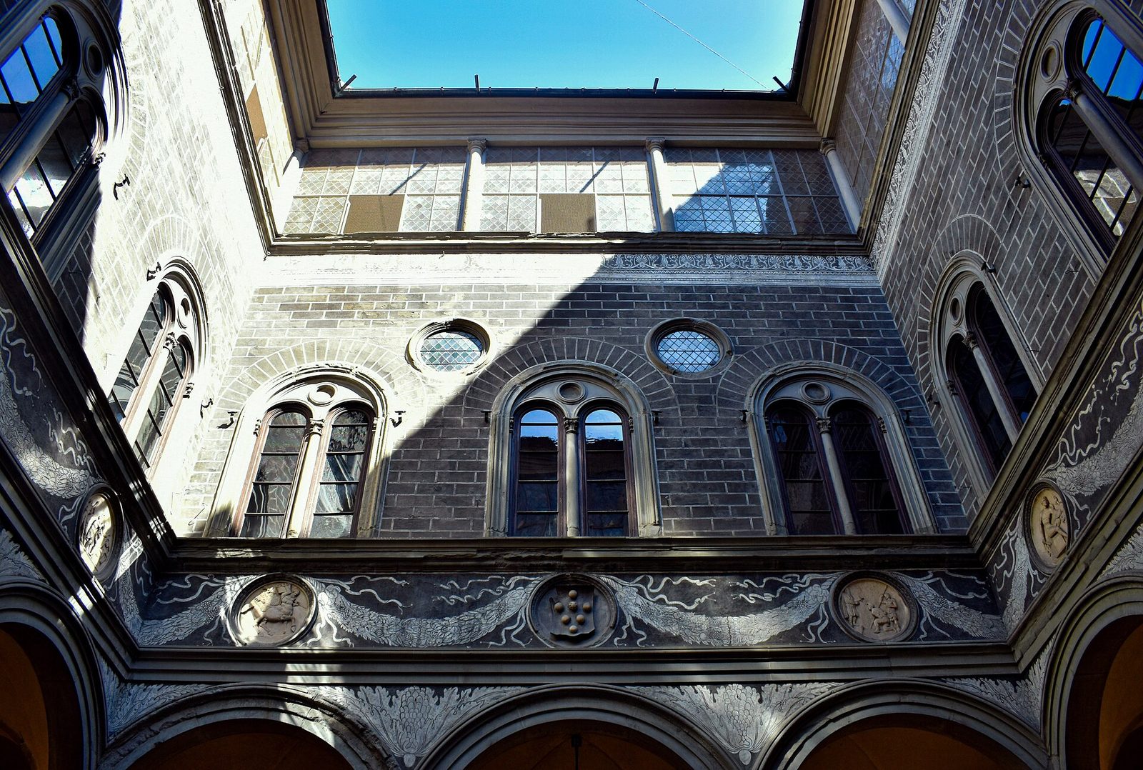 Looking up inside a square courtyard: an arcade on slender columns below, round-headed windows with slim mullions above, carved roundels in the frieze between them, and a strip of sky at the top.
