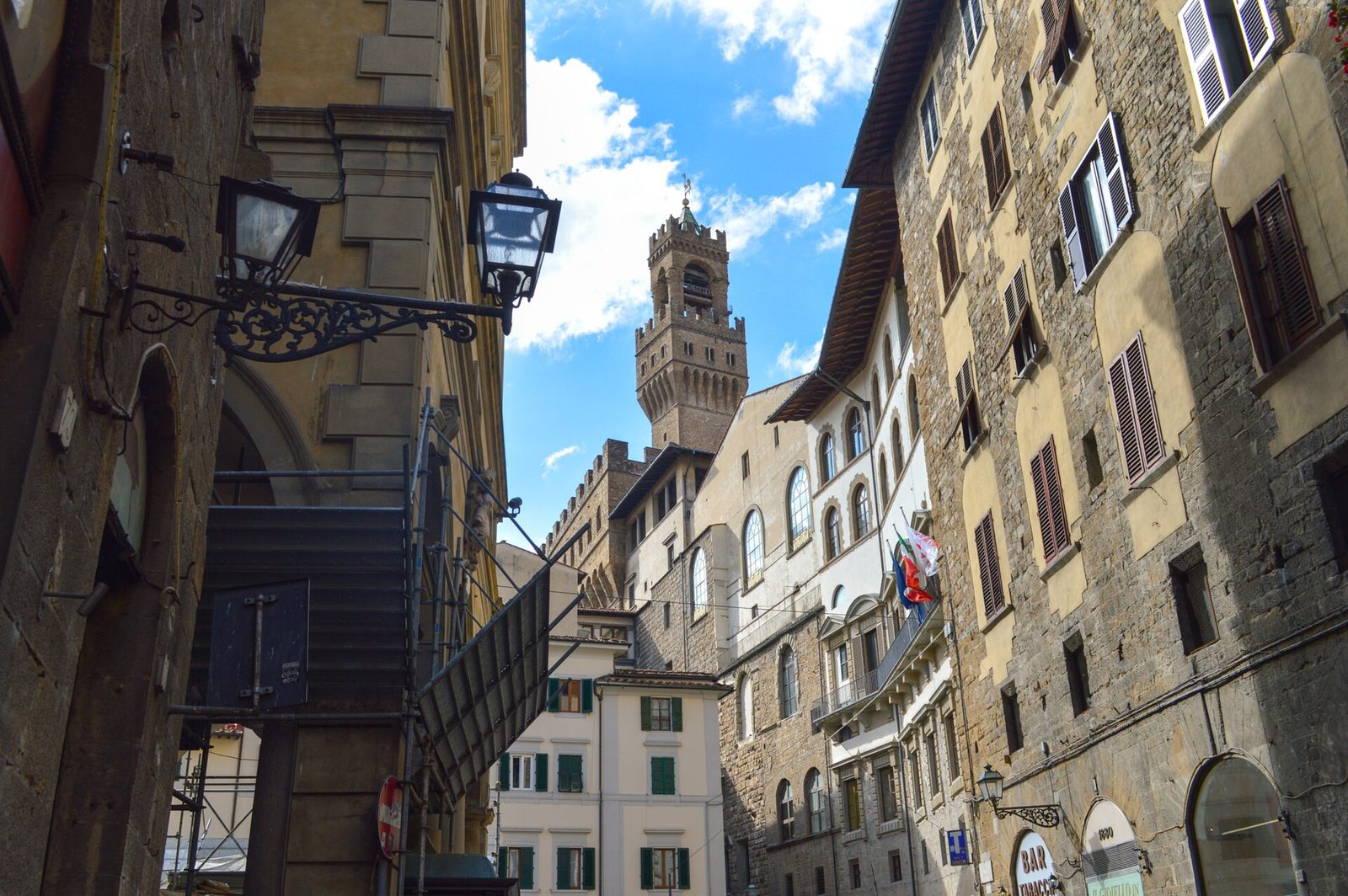 A narrow street of tall shuttered houses, with a square crenellated tower and its bell-stage rising above the rooftops at the end of the view.