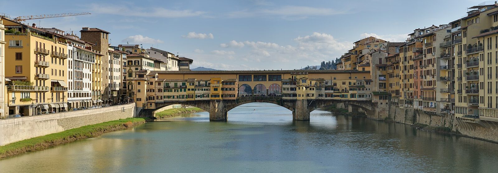 A low three-arched bridge spanning a green river, built over with shops and houses of several storeys, with a covered gallery running along the top and quays of stone buildings on both banks.
