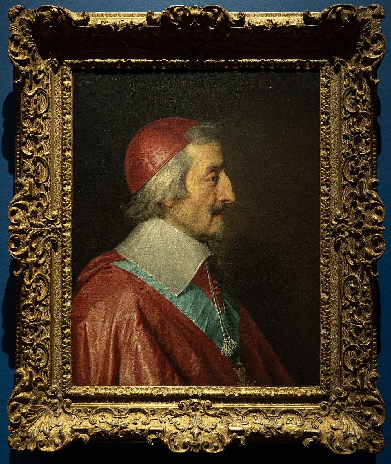 A full-length oil portrait of a man in a scarlet cardinal's robe and cape with a lace collar, standing with one hand extended, a heavy curtain and column behind him.