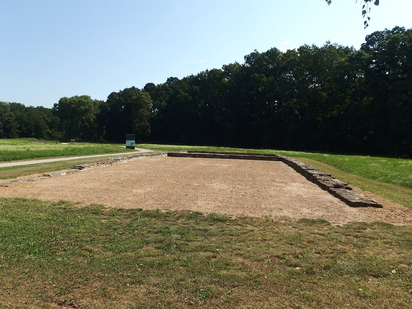 Low stone foundations of a long rectangular building laid out in a grassy clearing, woods behind.