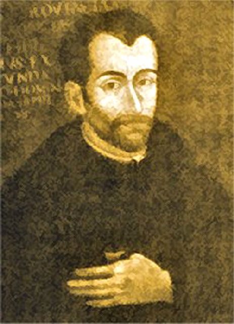 A sepia portrait of a bearded man in a dark cassock with one hand on his chest.