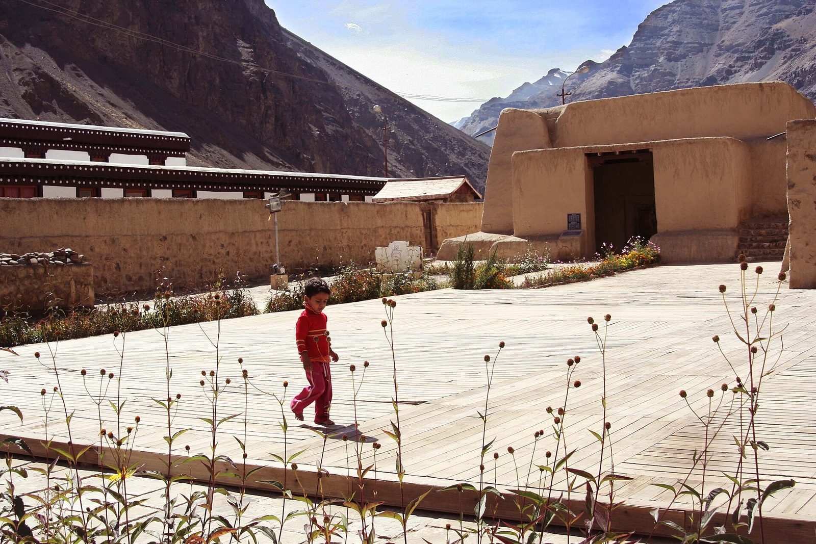 Mud-walled temple buildings around a paved courtyard under bare mountains, a child crossing it.