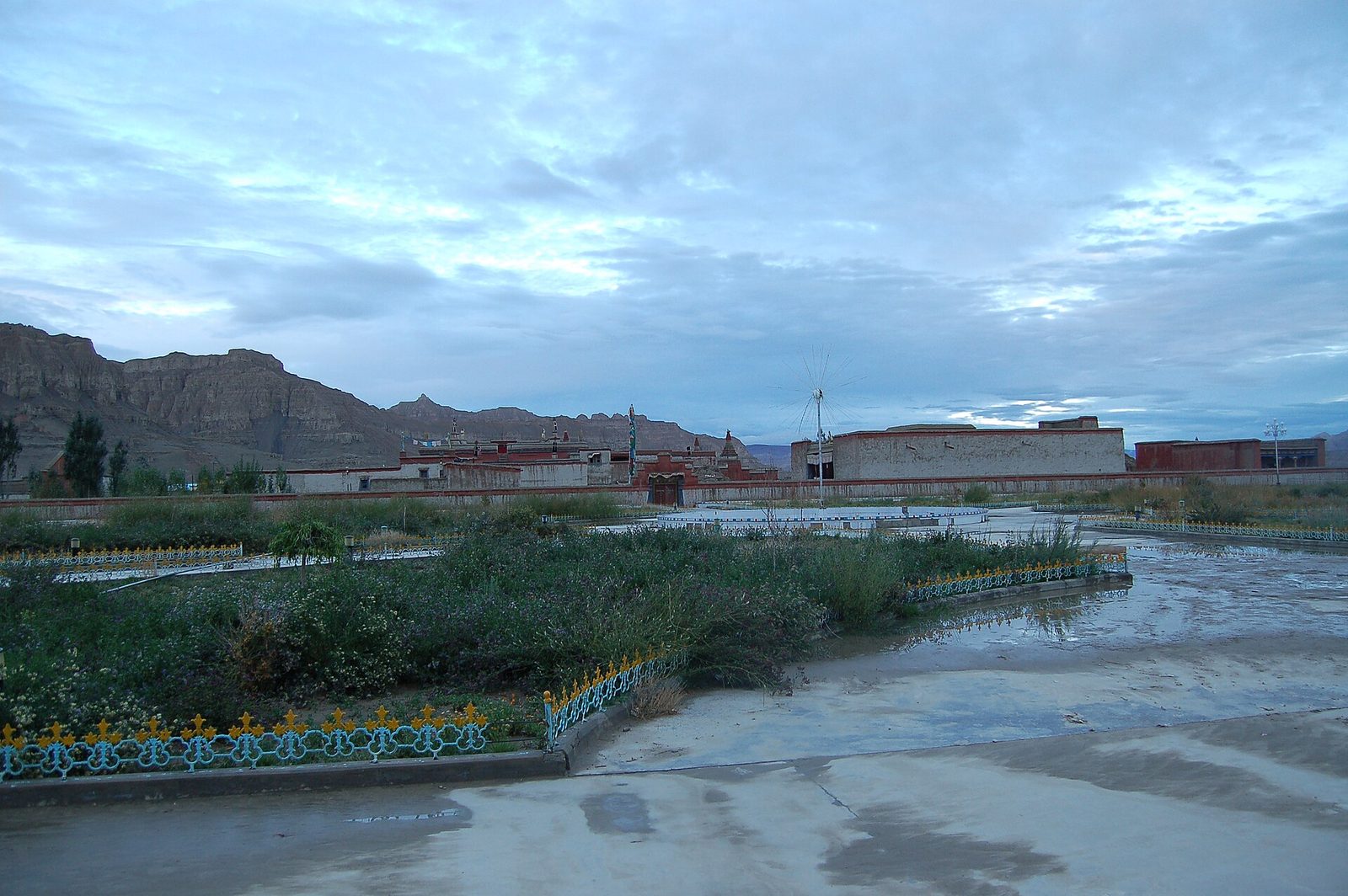 Low monastery buildings at the edge of a town under a cloudy evening sky, eroded hills behind.
