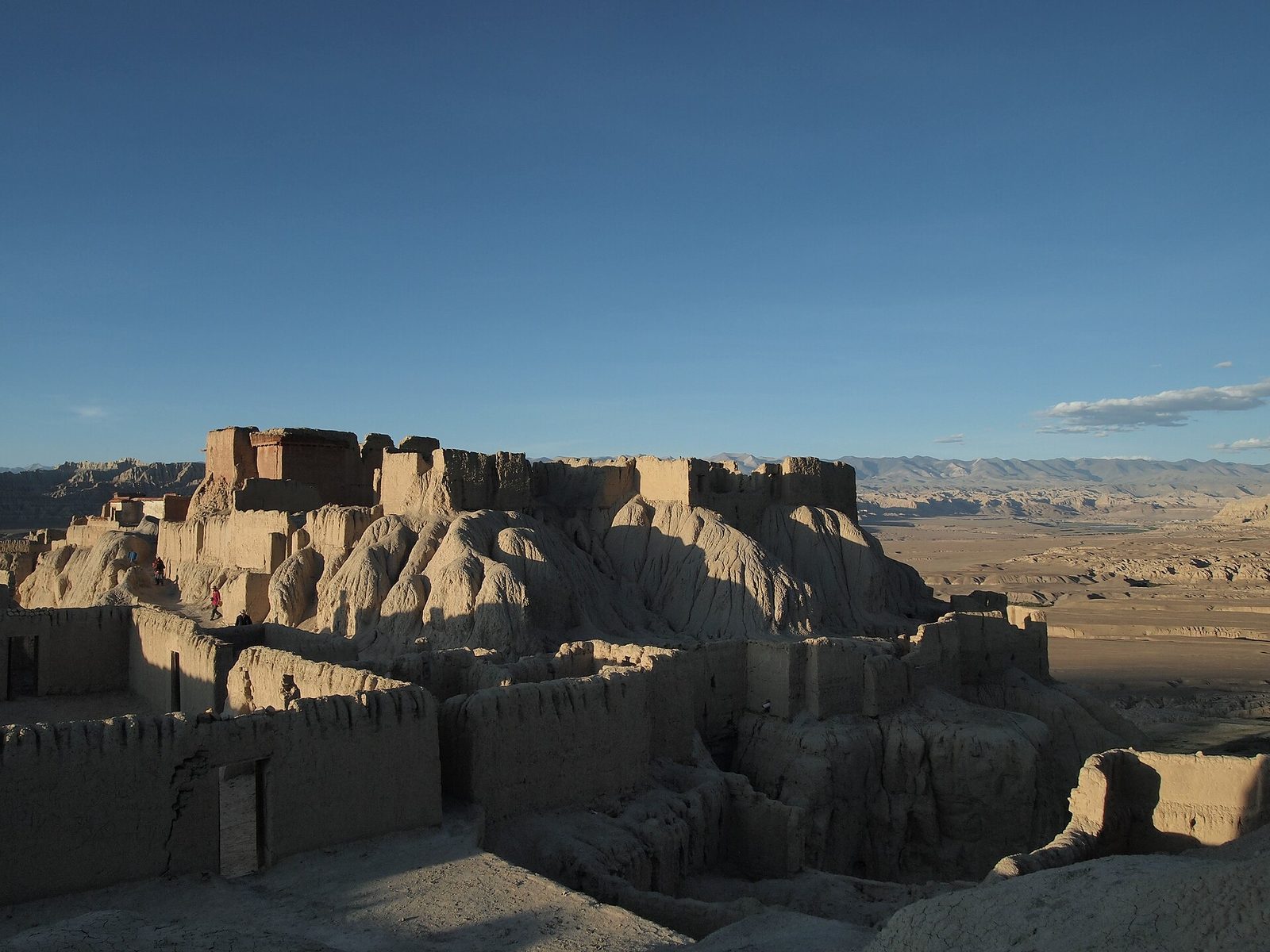 Ruined earthen buildings and walls climbing a steep eroded ridge above a wide valley at dusk.
