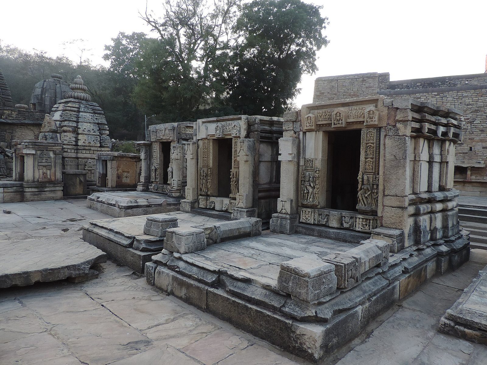 Rows of small stone shrines with carved doorways on a paved platform among trees.
