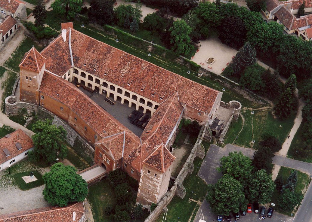 An aerial view of a castle with red roofs round a courtyard, among trees.