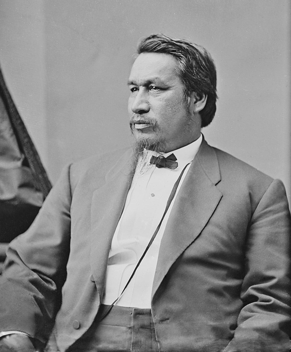 A photograph of a seated man in a dark suit and bow tie, looking to one side.