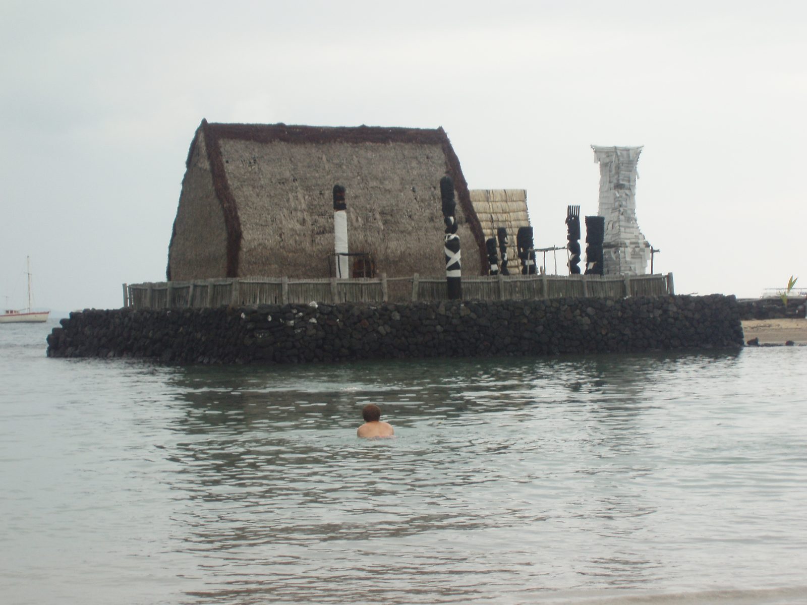 A thatched house and carved wooden images on a stone platform at the water's edge.