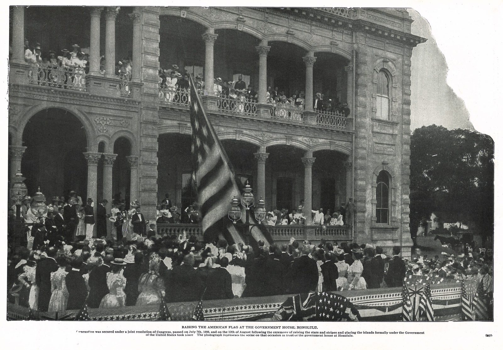 A halftone photograph of a crowd on the steps and balconies of a palace as a large flag is raised.