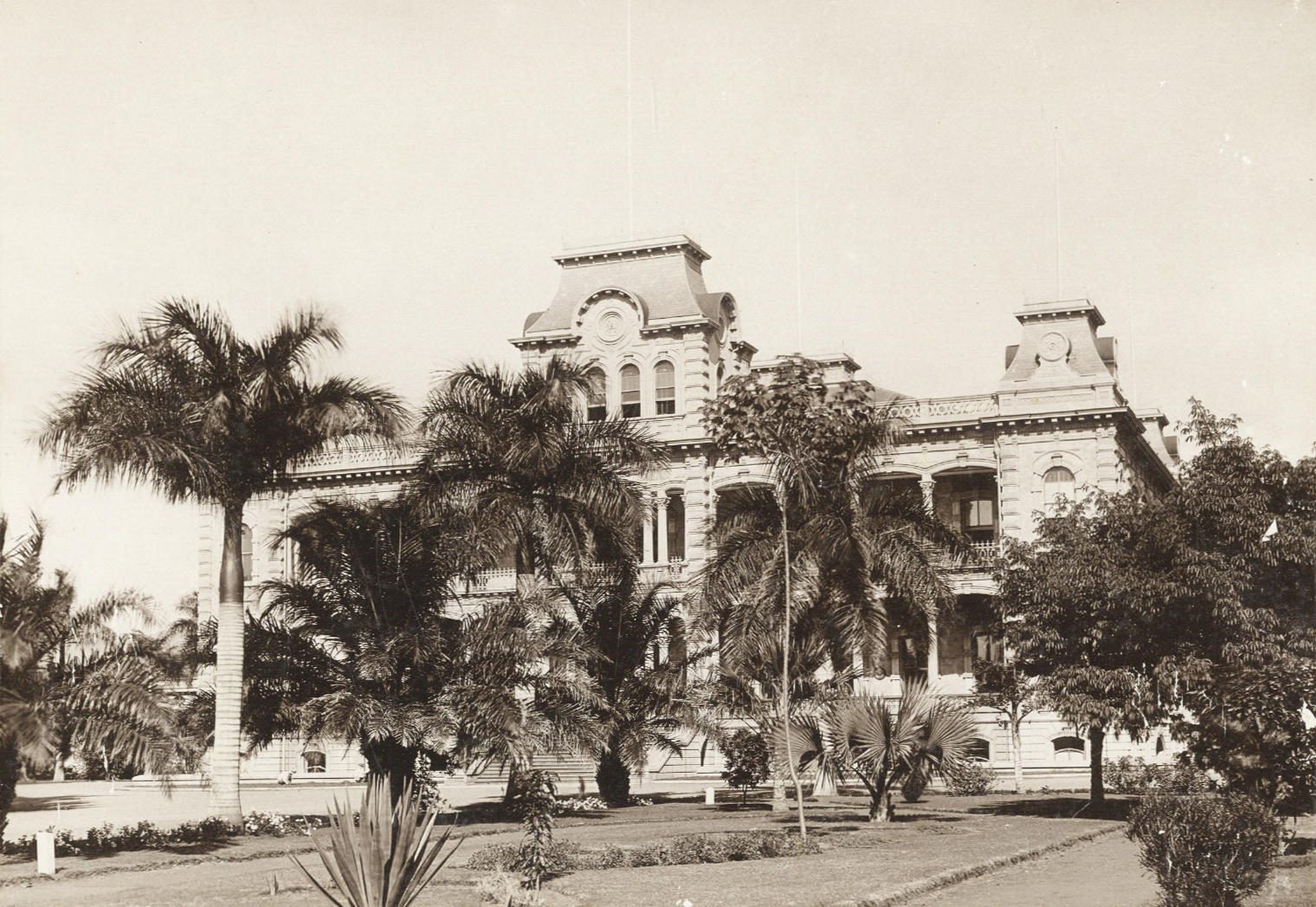 An old photograph of a two-storey palace with corner towers, behind palm trees and lawns.