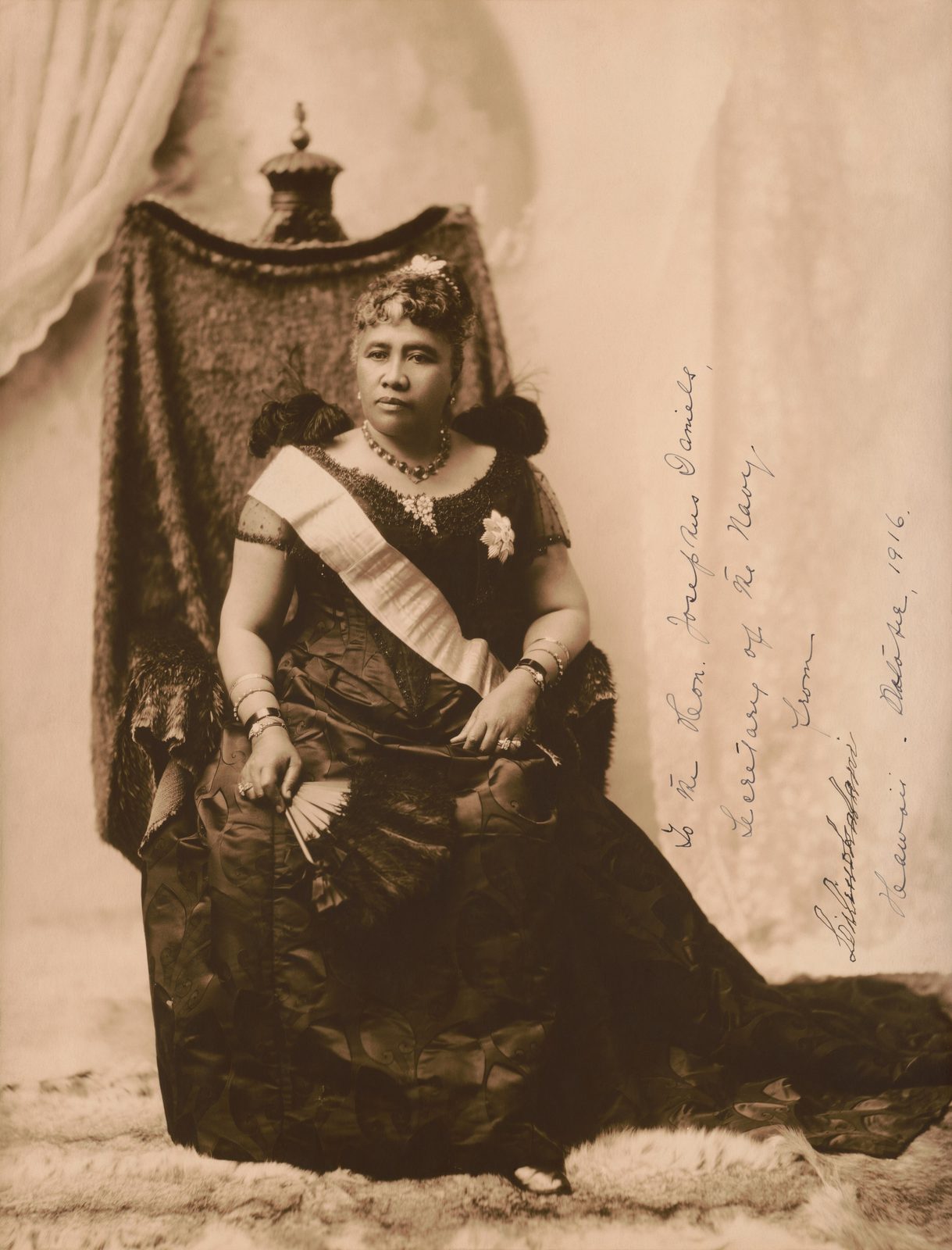 A sepia photograph of a woman seated on a carved chair in a dark gown and sash, signed in ink at the side.