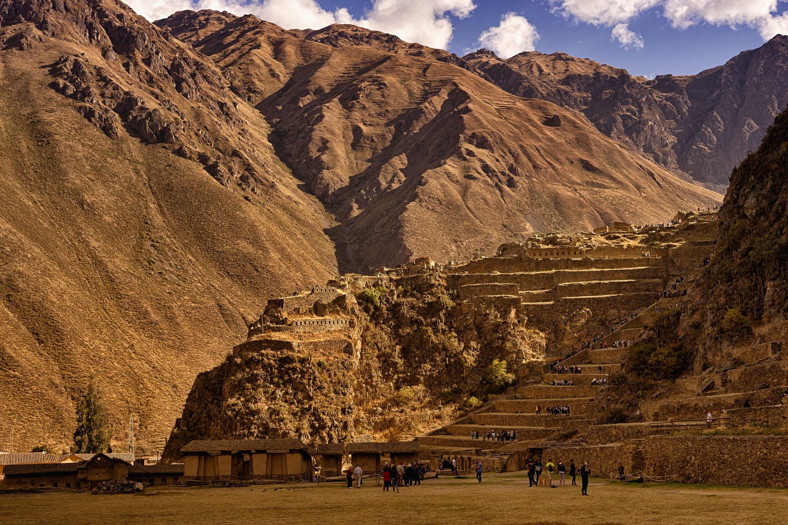 Stone terraces climbing a steep slope beneath mountains, a village below.