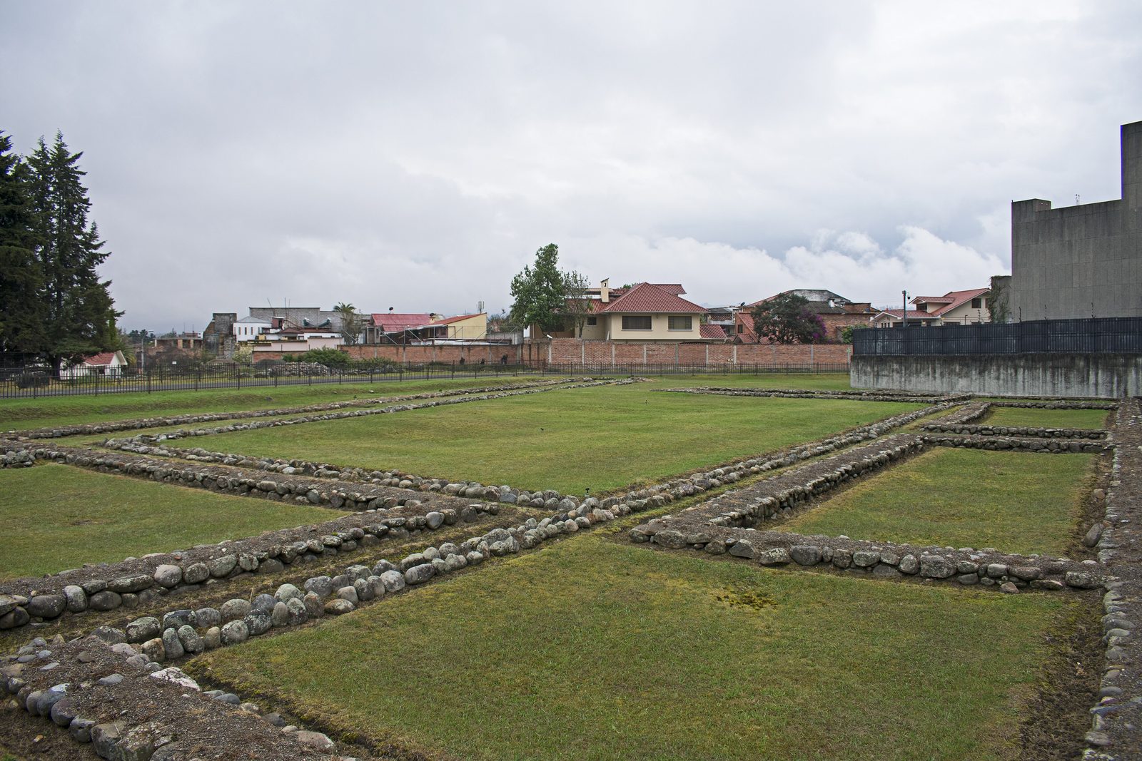 Low stone foundations of rectangular buildings in a lawn, with modern houses behind.