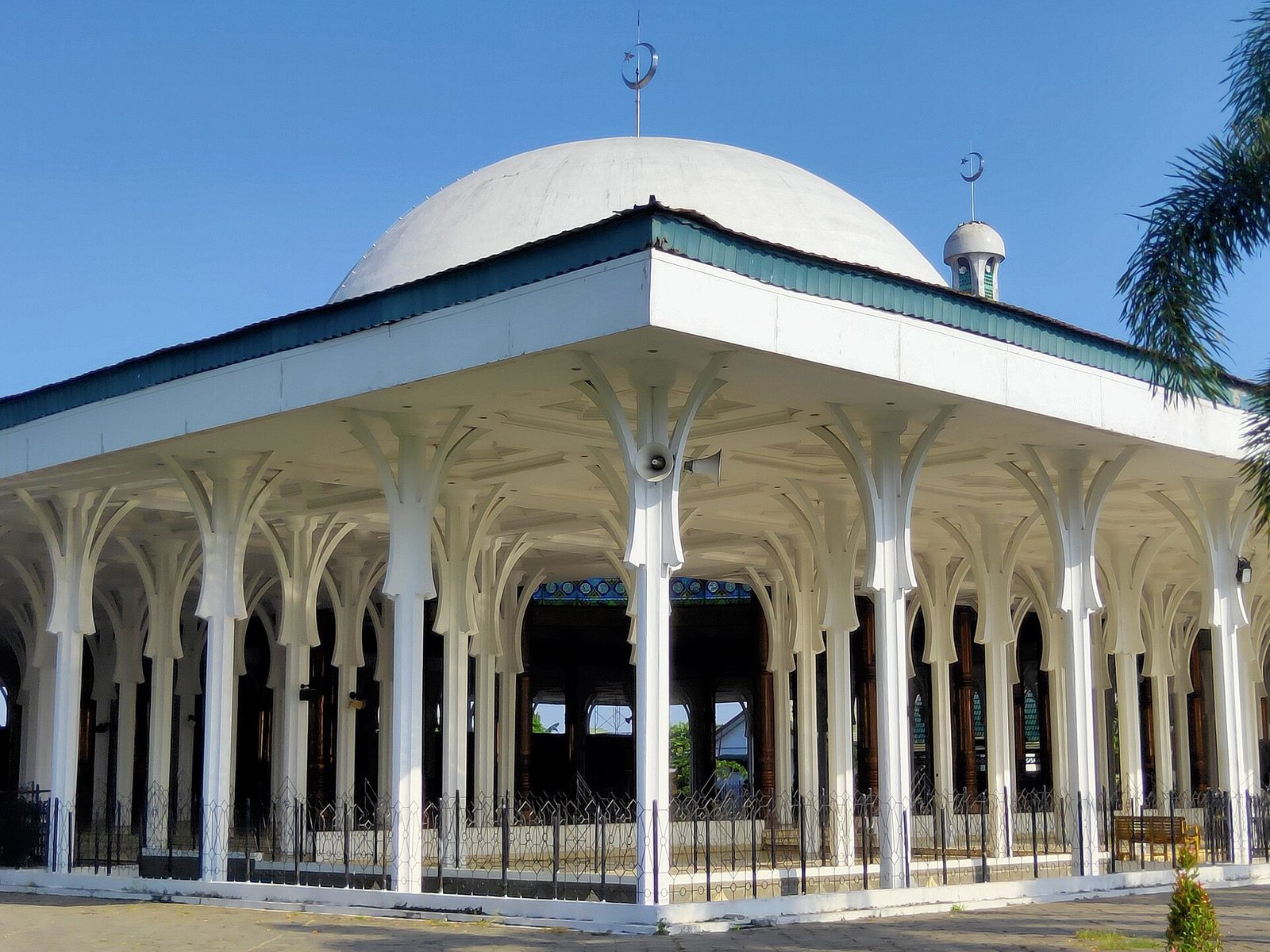 A large modern mosque with a wide shallow dome carried on rows of slender white columns, open on every side with no walls between them, standing on a paved terrace.