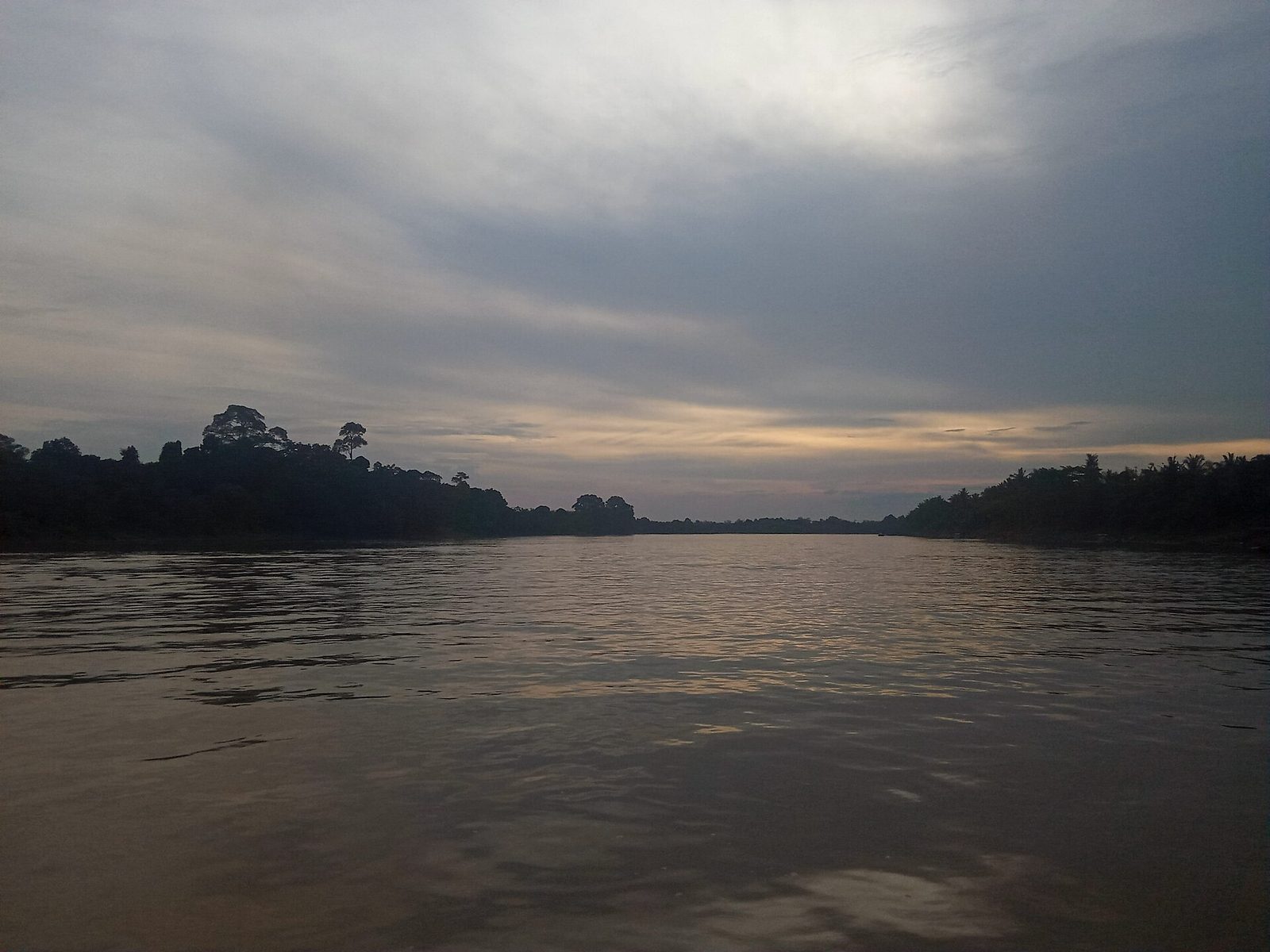 A wide river at dusk under a heavy sky, its surface flat and pale, with low forested banks running away on both sides to the horizon.