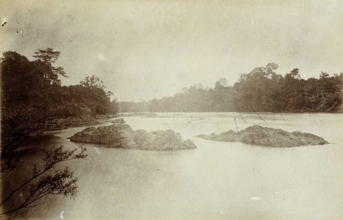 A sepia photograph looking up a broad river towards a wooded bend, with low rocky shoals breaking the surface in midstream and forest closing in on both banks.