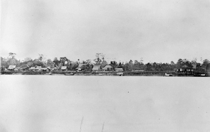 A sepia photograph of a riverside settlement: houses raised on posts along a low bank, palms and scrub behind them, and the water filling the foreground.