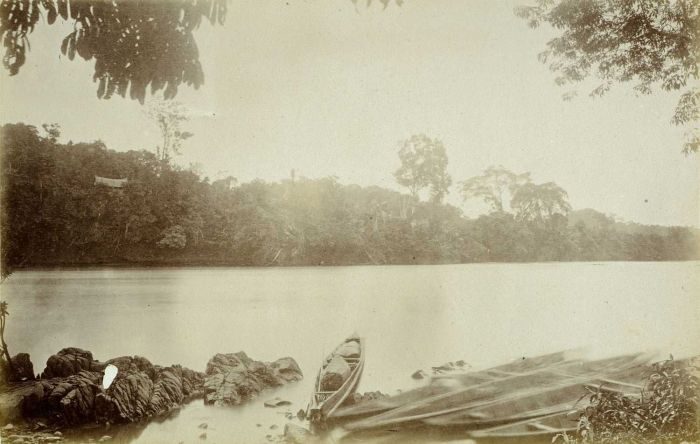 A sepia photograph of a river curving between wooded banks, with a long narrow boat drawn up on the shore in the foreground and a figure beside it.