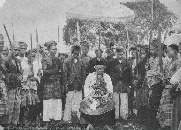 A photograph of a seated man in a dark cap and light jacket under a raised ceremonial parasol, surrounded by standing men in sarongs and headcloths holding upright spears, with trees behind.