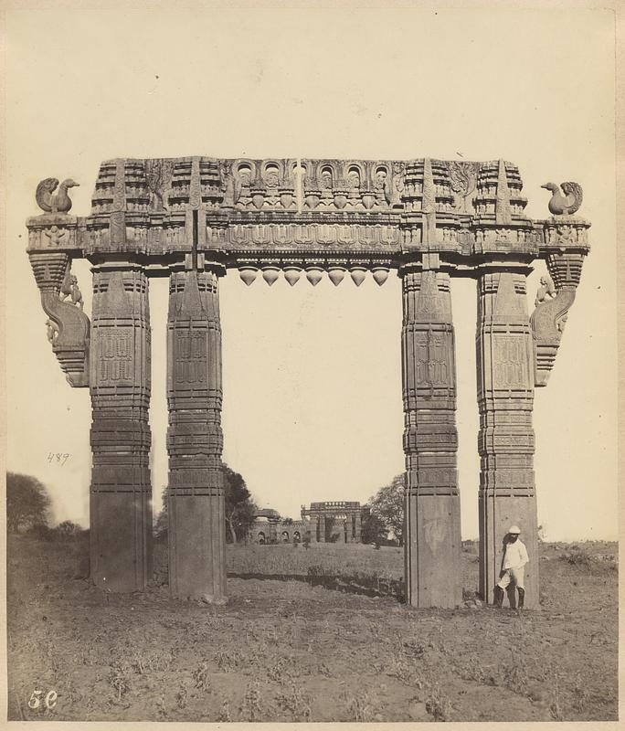 An old photograph of a tall carved stone gateway standing alone in open ground.