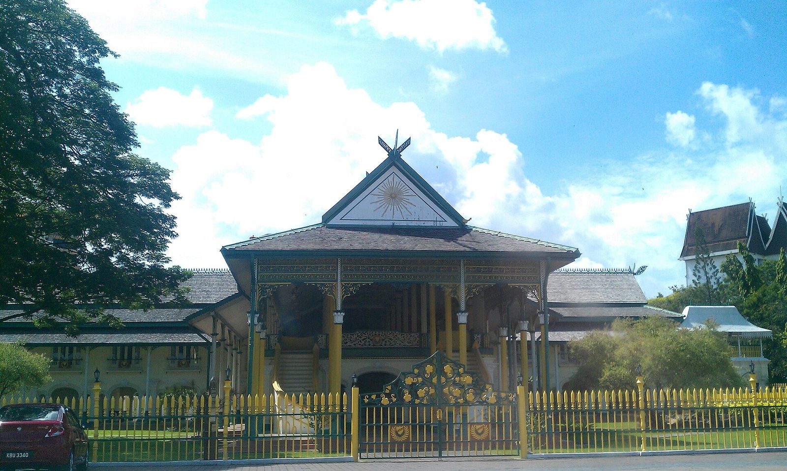 A long single-storey timber audience hall with a steep tiered roof and a crested ridge, standing behind a yellow and black railing on a paved forecourt.