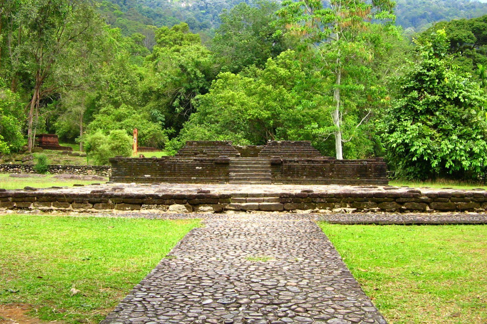 A low rectangular platform of dressed stone blocks standing on mown grass, a cobbled path leading up to its steps, with dense forest and a hillside rising behind.