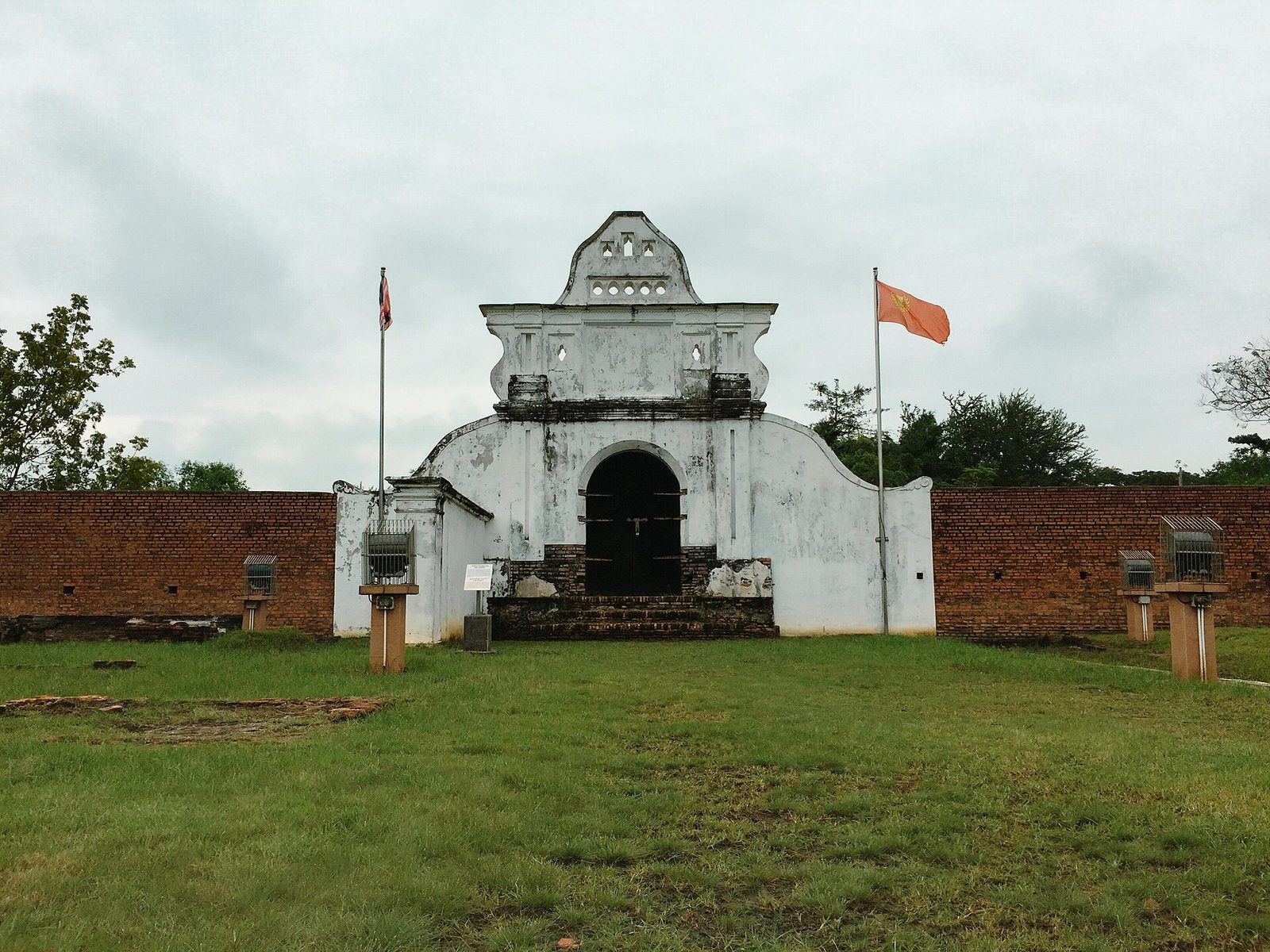 A whitewashed gateway with a scrolled gable and a barred arch, set in a long wall of weathered brick, with flagpoles on either side and mown grass in front.