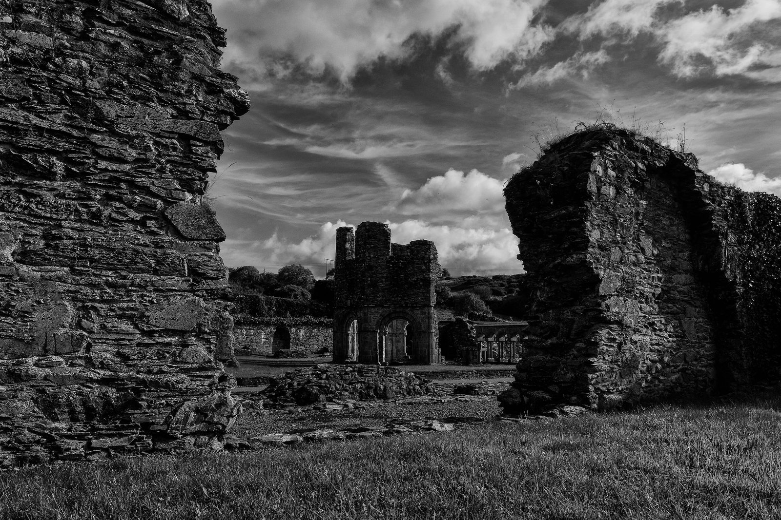 A black-and-white photograph of ruined abbey walls framing more ruins under a cloudy sky.