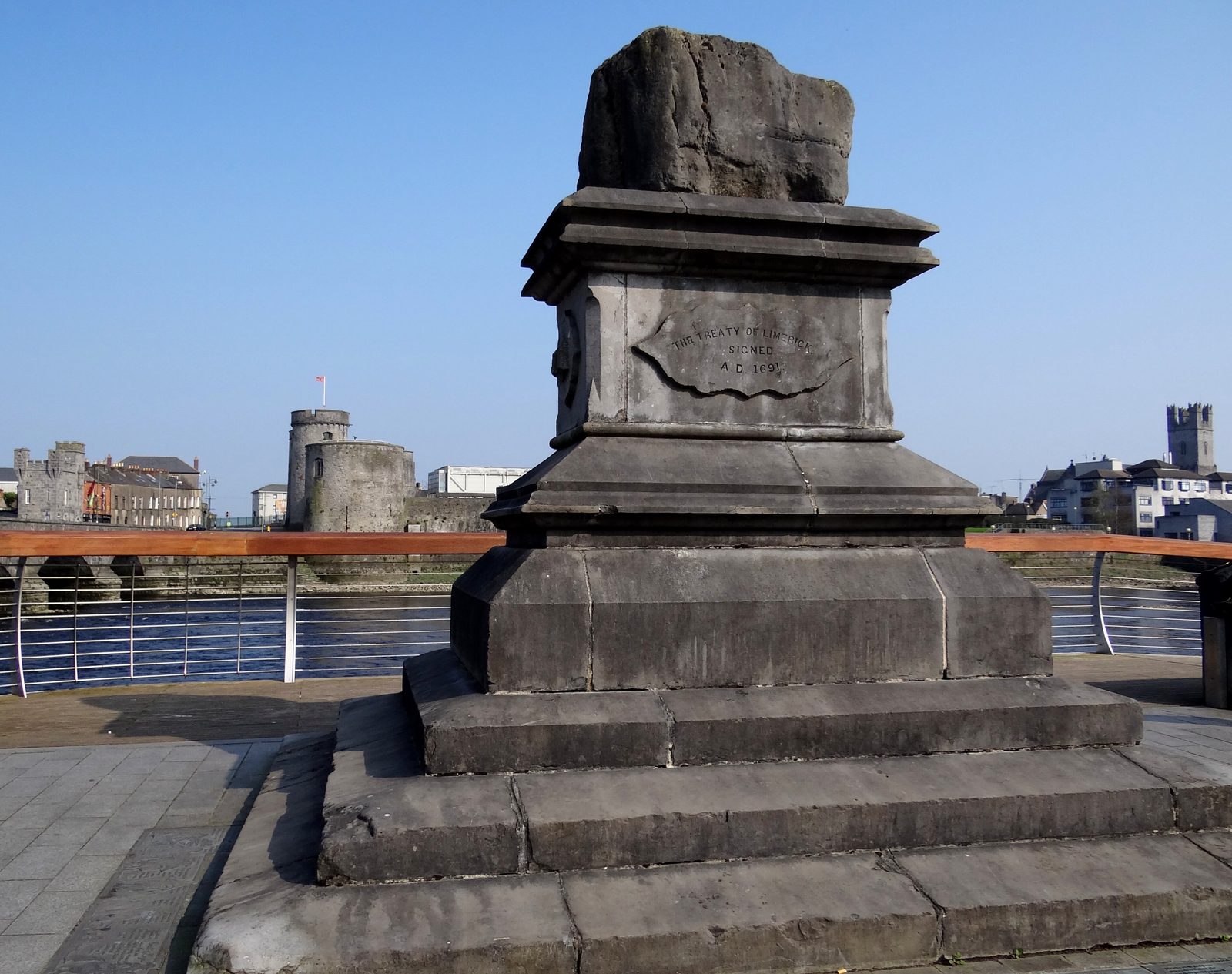 A rough block of stone on a stepped pedestal beside a river, a castle across the water.