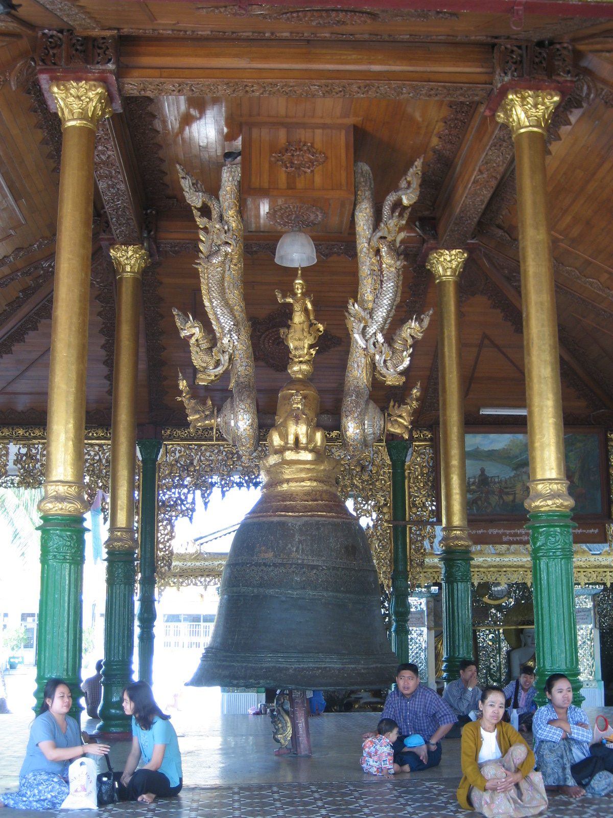 A large bronze bell hanging in an ornate gilded pavilion, with people seated on the floor beside it.