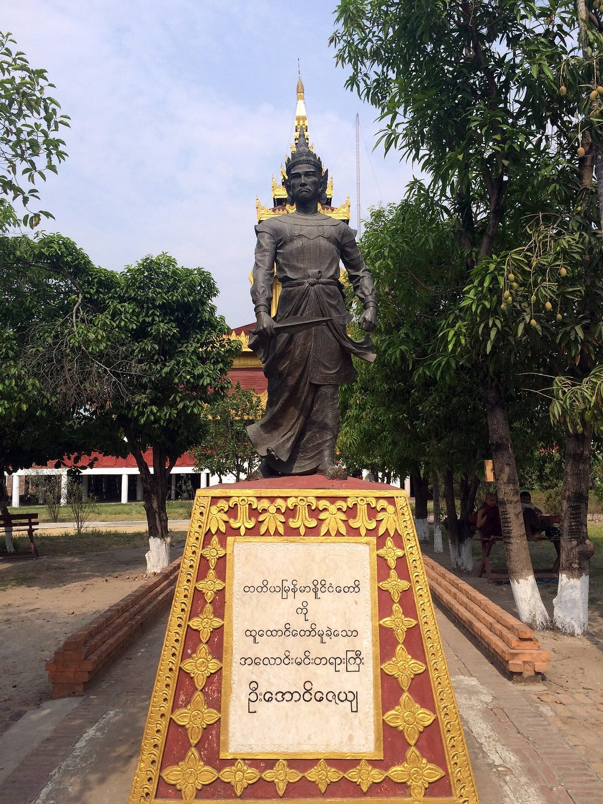 A modern standing statue of a crowned king on a red and gold pedestal inscribed in Burmese, among trees.