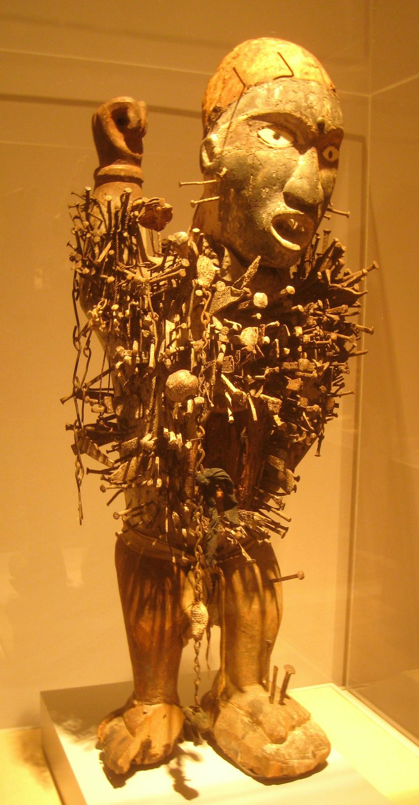 A standing wooden figure with one raised arm, its body bristling with driven nails and iron blades.