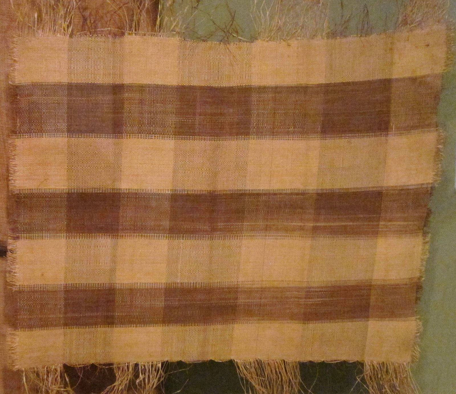 A square of plain-woven palm-fibre cloth in brown and buff checks with a fringed edge.