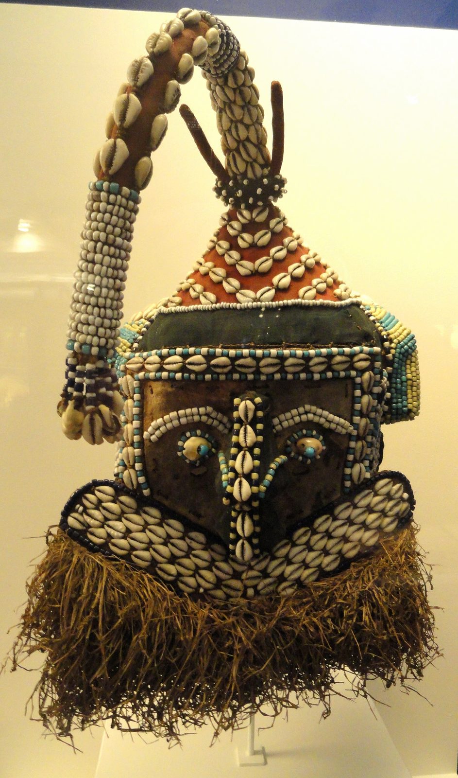 A helmet mask covered in cowrie shells and beads, with a long beaded trunk rising from the top and a fringe of fibre below.