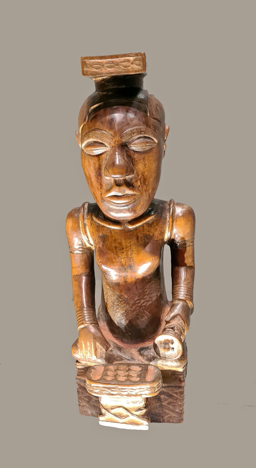 A seated carved wooden figure of a king in a flat cap, a game board with rows of cups on the base before him.