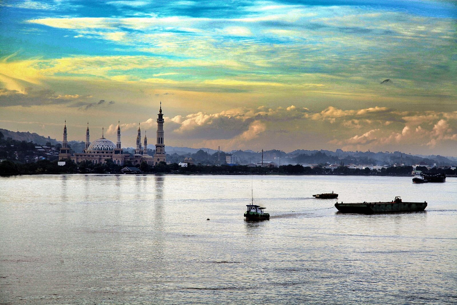 A wide river at sunset, small boats and a barge on the water and a low town with a domed mosque and slender minarets along the far bank.