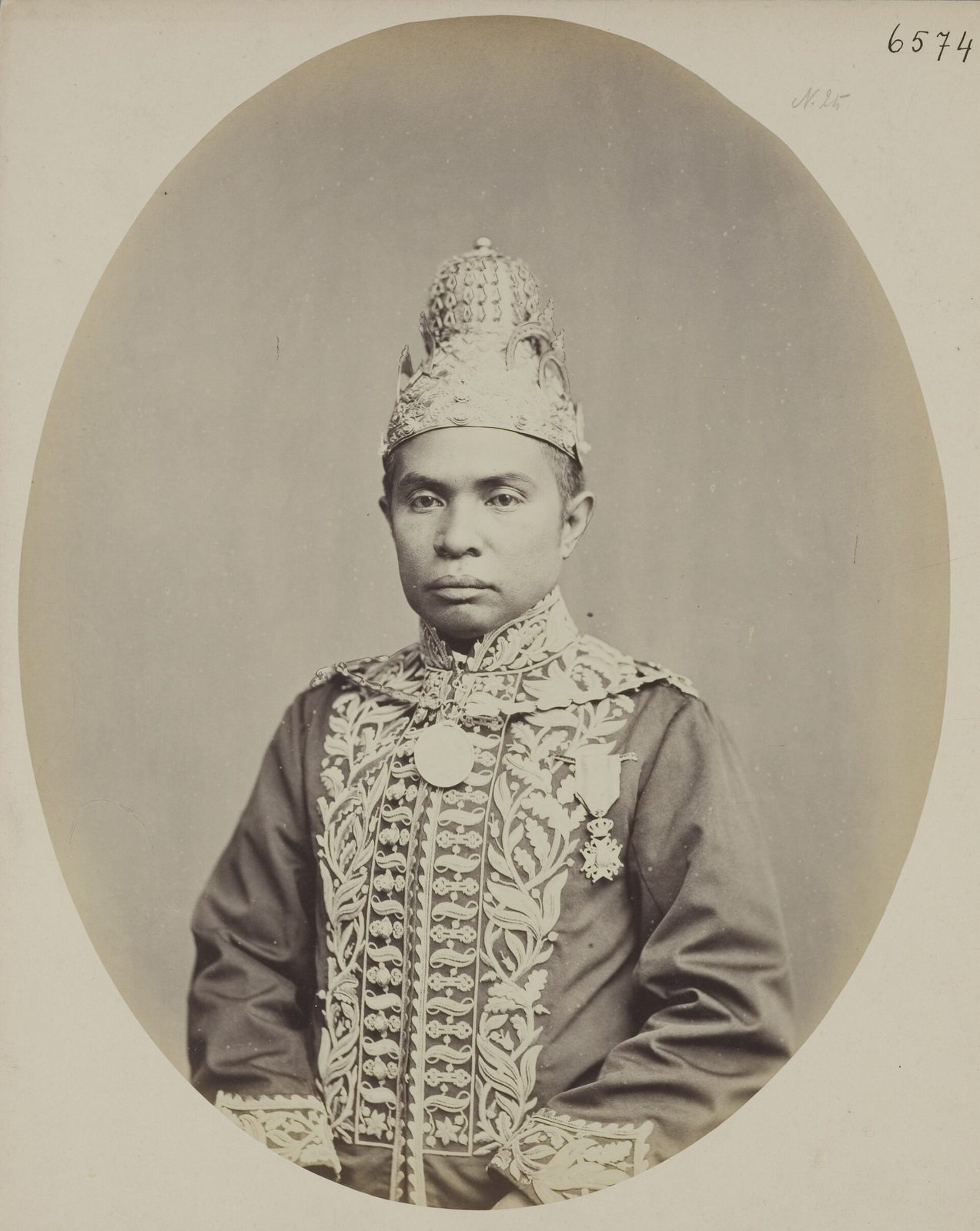 An oval studio photograph of a seated man in an embroidered jacket and a tall ornamented cap, looking directly at the camera.