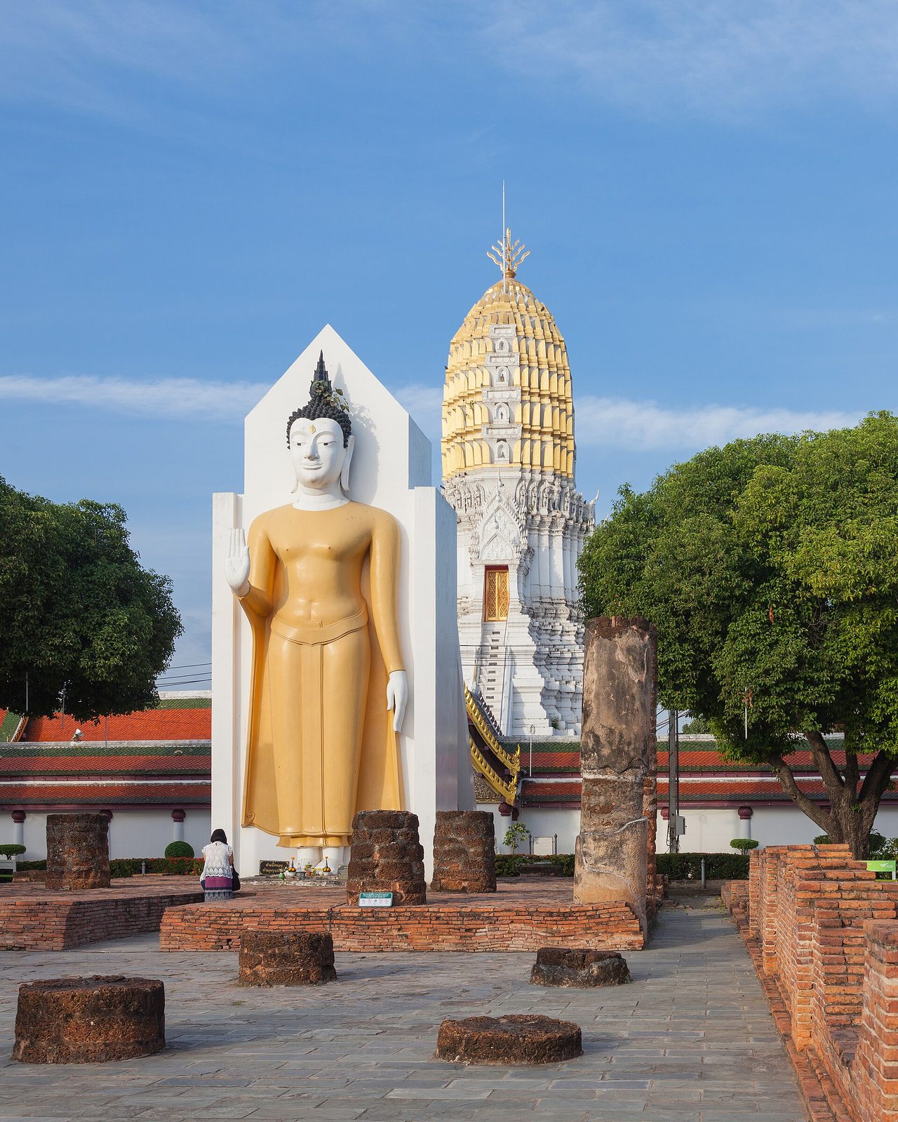 A standing gilded Buddha beside a white tower among ruins.